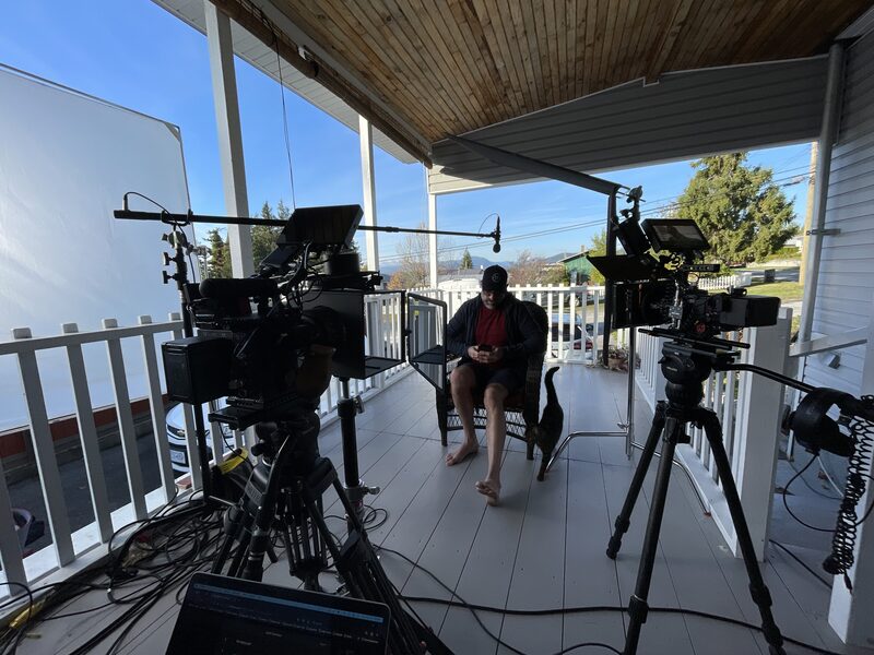 Man sitting on a porch with a black cat, surrounded by filming equipment and cameras, with a white fence, trees, and houses in the background.