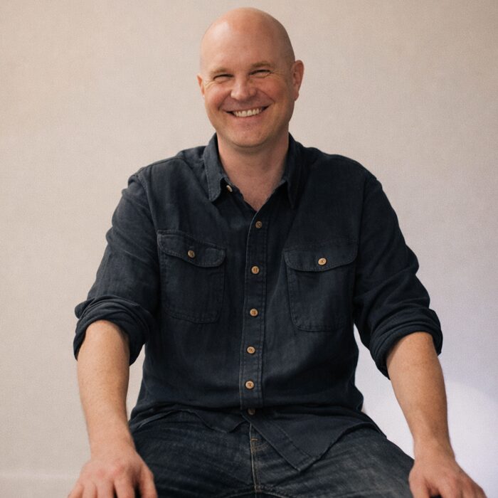 A smiling man with a bald head, wearing a black button-up shirt with rolled-up sleeves, sitting cross-legged on a white chair against a plain beige wall.