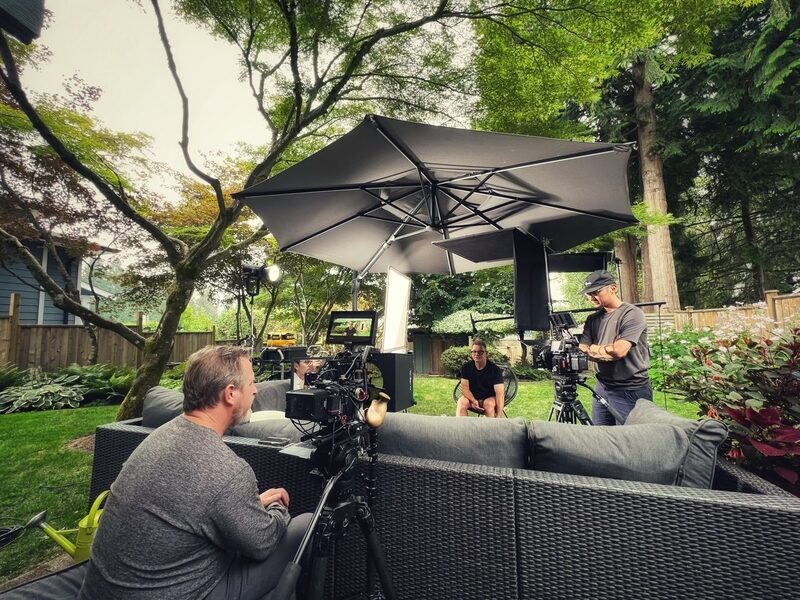 Filming crew recording a scene in a backyard with greenery, trees, and outdoor furniture, using professional cameras and lighting equipment.