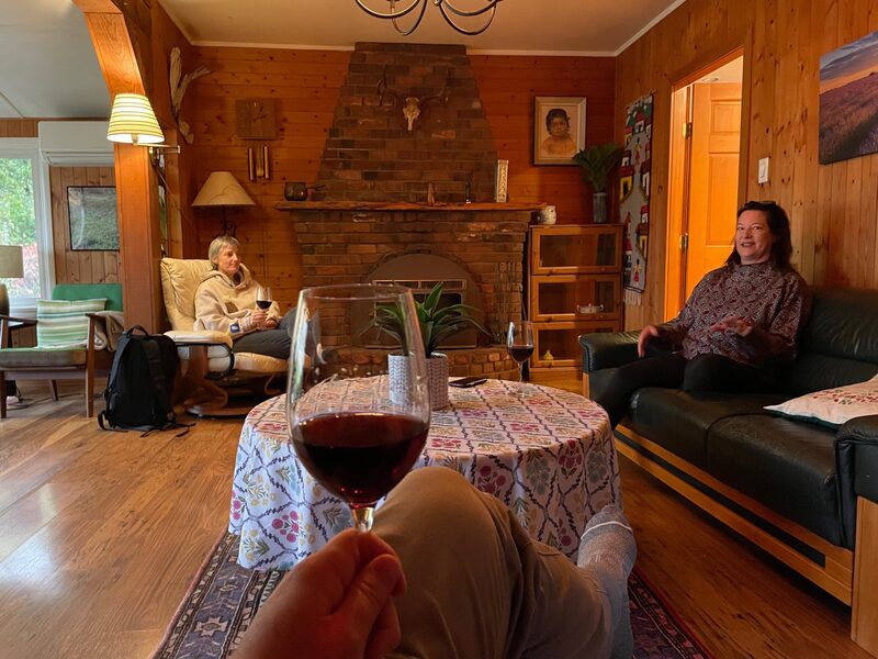 A person holding a glass of red wine while sitting on a couch, with two women in the background in a cozy wood-paneled living room, one sitting on a cushioned chair and the other on a sofa, near a brick fireplace.
