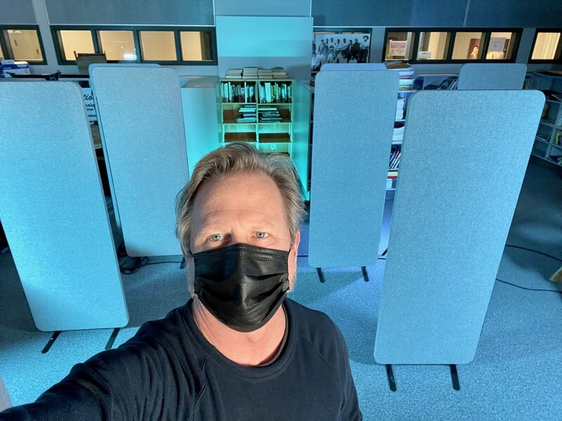 A man wearing a black face mask and a black shirt taking a selfie in a room with blue sound barriers and a bookshelf in the background.