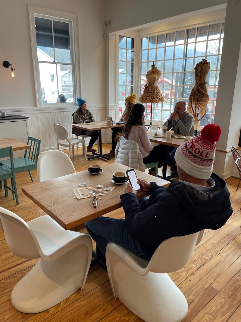 People sitting and chatting in a cozy cafe with large windows and decorative art pieces, wearing winter hats and jackets.
