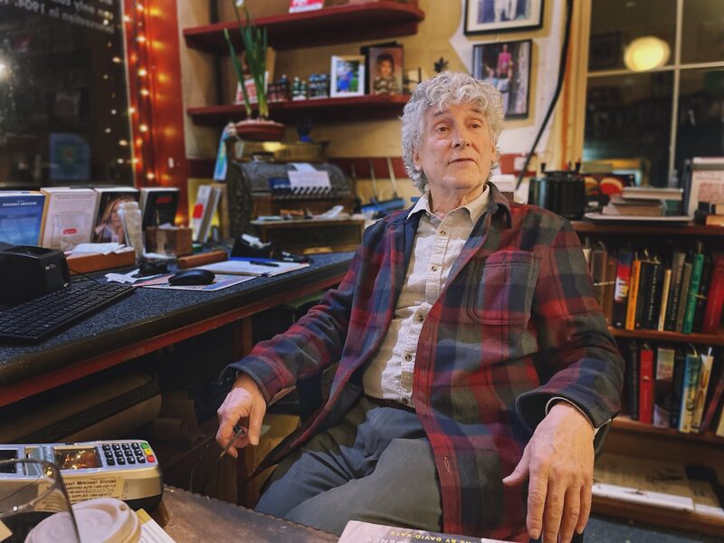 An elderly man with curly gray hair sits at a cluttered desk in a cozy bookstore or library. He is wearing a tan shirt and a red and dark plaid jacket. Behind him, bookshelves filled with books, framed photographs, and various decorative items are vi