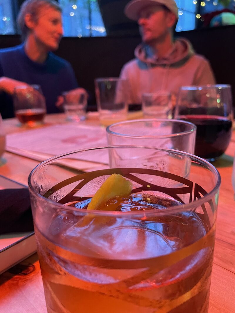 Close-up of a glass of iced tea with a lemon wedge on top, on a wooden table at a restaurant. In the background, two people are sitting and talking, slightly out of focus.