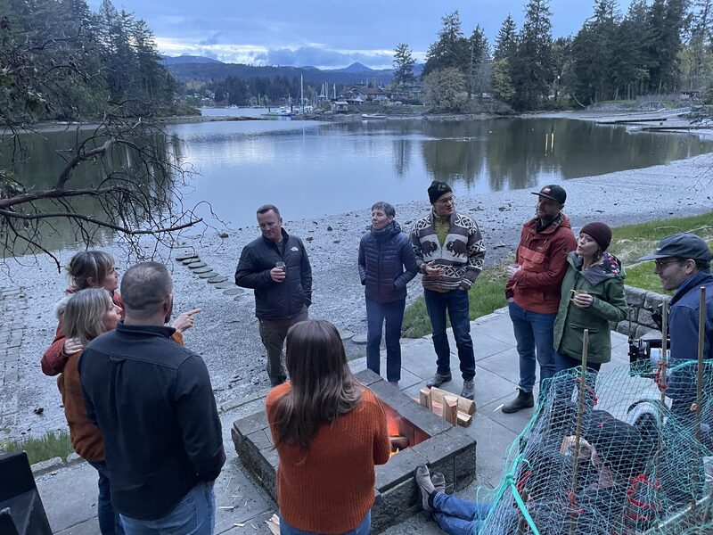 Group of people gathered around a fire pit on a patio near a lake, with trees, boats, and mountains in the background during evening or dusk.