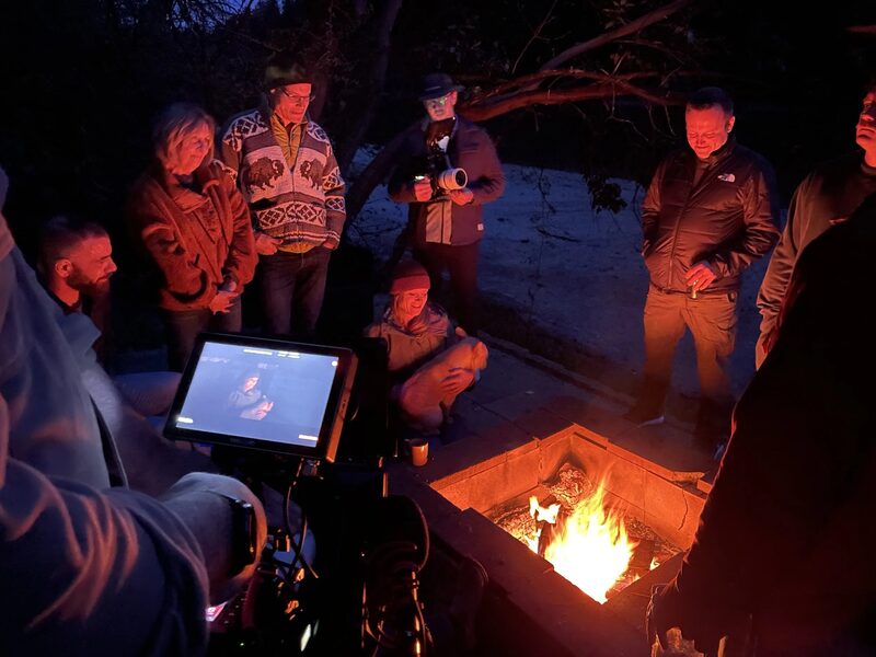 Group of people gathered around a campfire outdoors at night, some standing and some sitting, with a person holding a camera taking a photo.