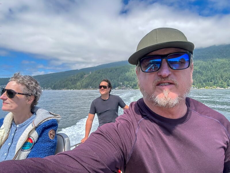 Three people on a boat with a mountain and forested shoreline in the background, enjoying a clear day with some clouds.