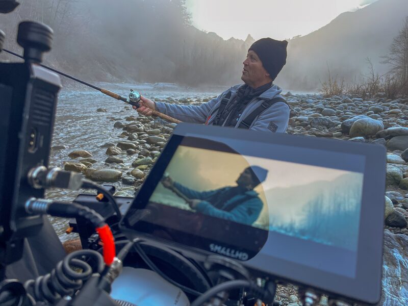 A man fishing in a rocky river with mountains in the background, seen through a camera monitor and equipment.