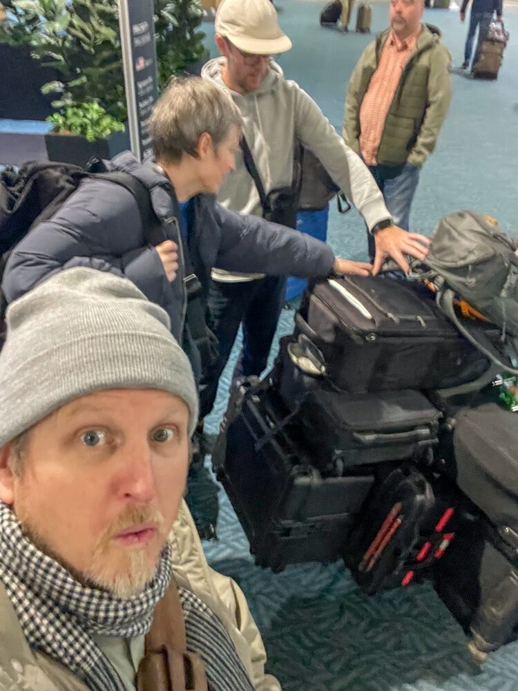 A man taking a selfie in an airport with three travelers behind him, all with luggage. The travelers are looking at the luggage cart, and one woman is reaching out to touch it. The man in the foreground wears a gray beanie, patterned scarf, and beige