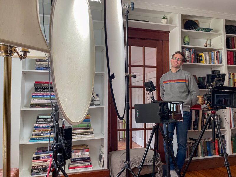 A man stands in front of a camera setup in a cozy, well-lit room with bookshelves, smiling and wearing glasses and a gray sweater with rainbow stripes.