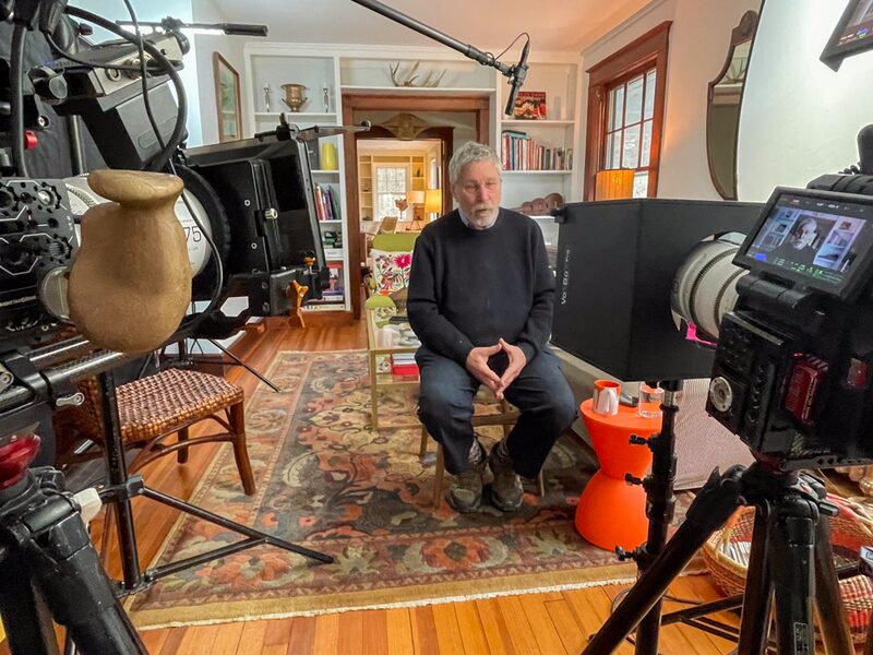 Older man with gray hair and beard sitting on a chair in a warmly lit living room with wooden floors and furniture, being filmed with professional camera equipment.