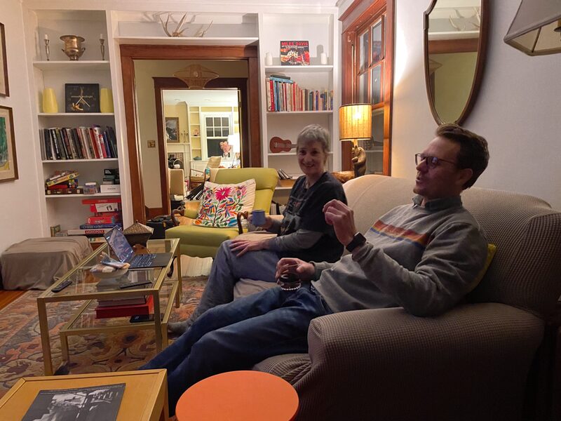 Two people sitting on a couch in a cozy living room, one holding a mug and the other with a glass, near a coffee table with a laptop and books, with bookshelves, a lamp, and pictures on the wall in the background.