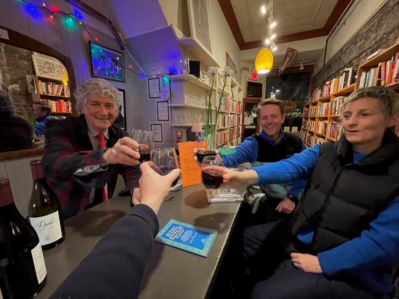 Four people in a cozy bookstore or cafe raising glasses of red wine for a toast, smiling, with bookshelves, string lights, and a mirror in the background.