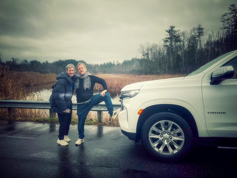 Two smiling women stand beside a white SUV near a swampy wetland on a cloudy day, with trees in the background.
