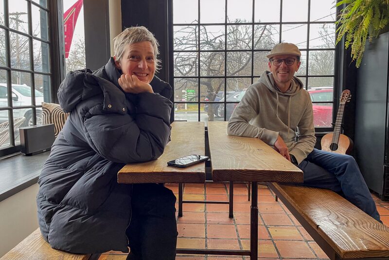 A smiling woman with short gray hair wearing a black coat sits at a wooden table in a coffee shop. A man with glasses, a beige cap, and a gray hoodie sits on the other side of the table, with an acoustic guitar on the floor next to him. The backgroun
