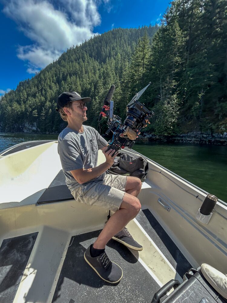 A man operating a camera on a boat in a scenic river with forested mountains and a partly cloudy sky.