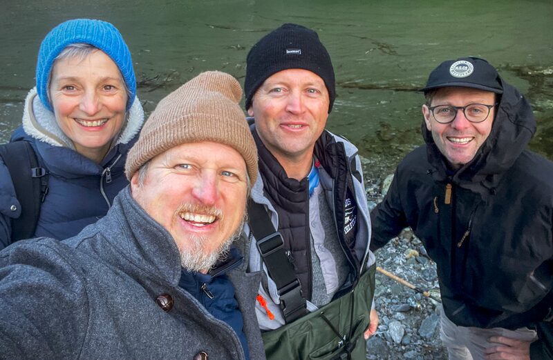Four adults outdoors near a river, smiling and wearing jackets and hats.