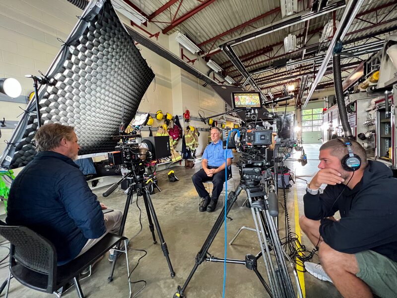A man in a blue uniform being interviewed or filmed in a fire station's garage, surrounded by film equipment and firefighters' gear hanging on the wall.