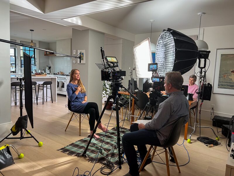 A woman is sitting on a chair in a home kitchen, being filmed during an interview or video recording. The setup includes multiple professional lighting and audio equipment, with a crew member sitting nearby observing. The kitchen has a modern design