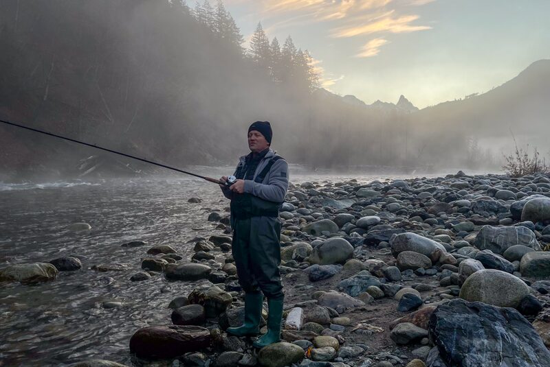 A person fishing with a fishing rod at a rocky riverbank during dawn or dusk, with mist rising from the water and mountains in the background.