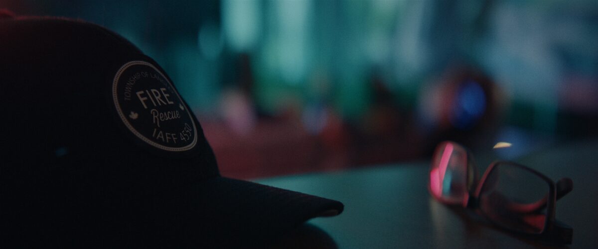 A black firefighter's cap with a patch reading 'Township of Lancaster Fire Rescue IAFF 4550' and a pair of glasses on a surface in a dimly lit room.