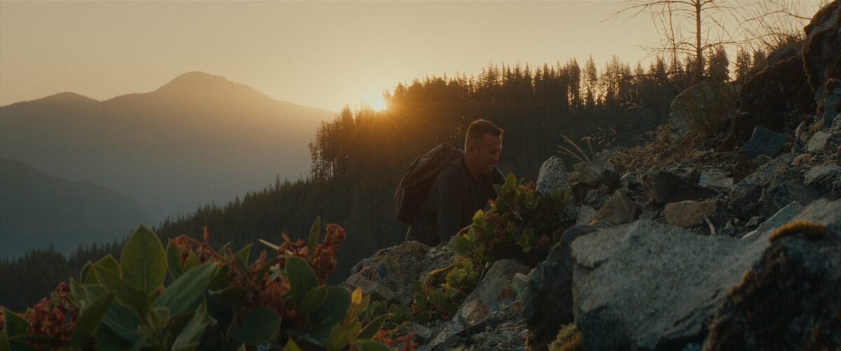 A person hiking uphill on a rocky trail at sunset, with mountainous terrain and trees in the background.