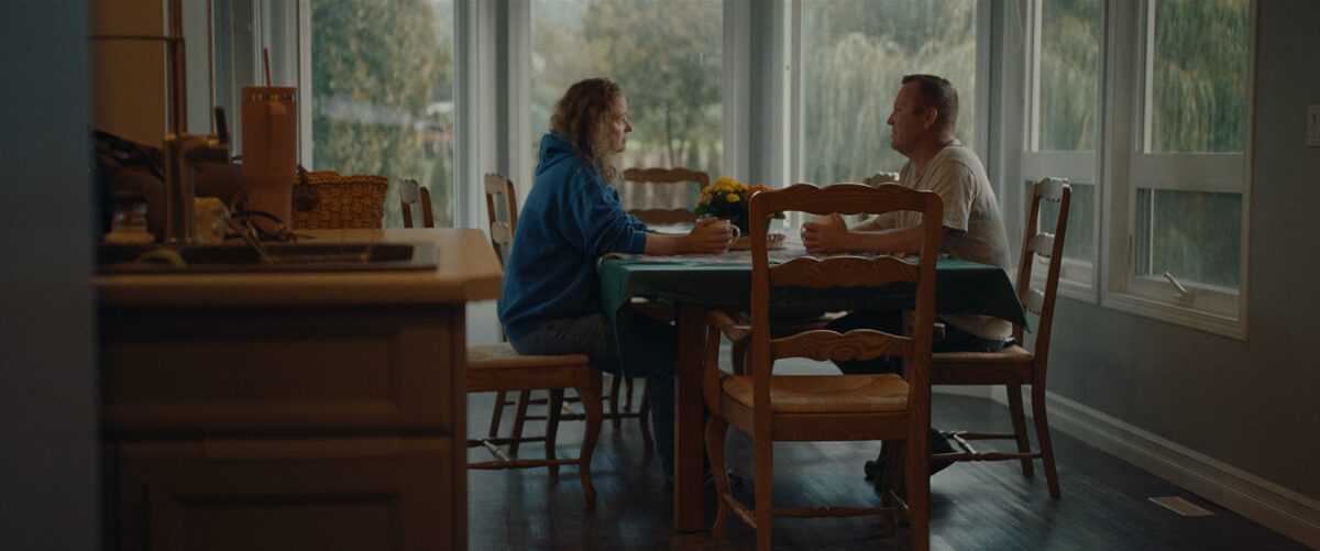 A woman and a man sitting across from each other at a dining table, engaged in conversation, in a sunlit kitchen with large windows showing a view of trees outside.