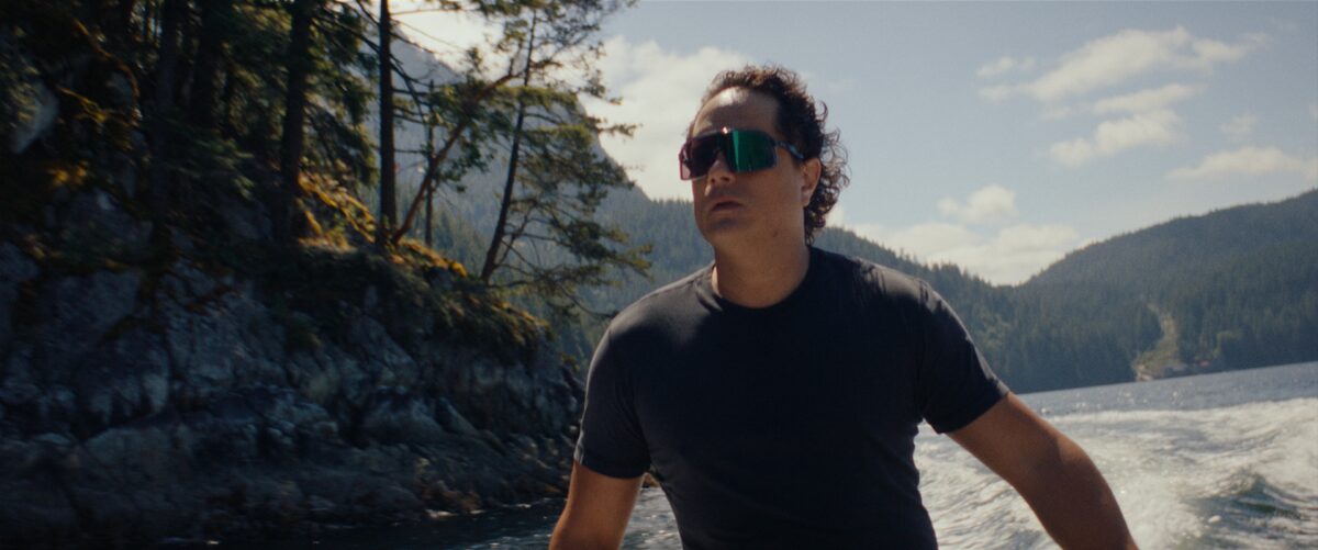 A man wearing sunglasses in a black t-shirt on a boat in a lake surrounded by mountains and trees on a sunny day.