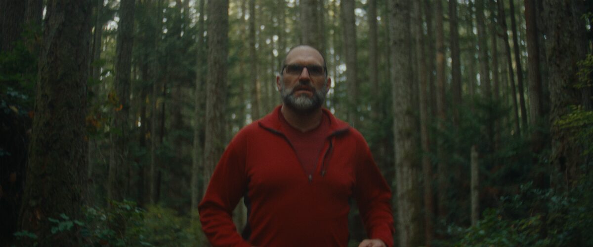 A man with glasses, gray beard, and red jacket running through a forest with tall trees and dense foliage.