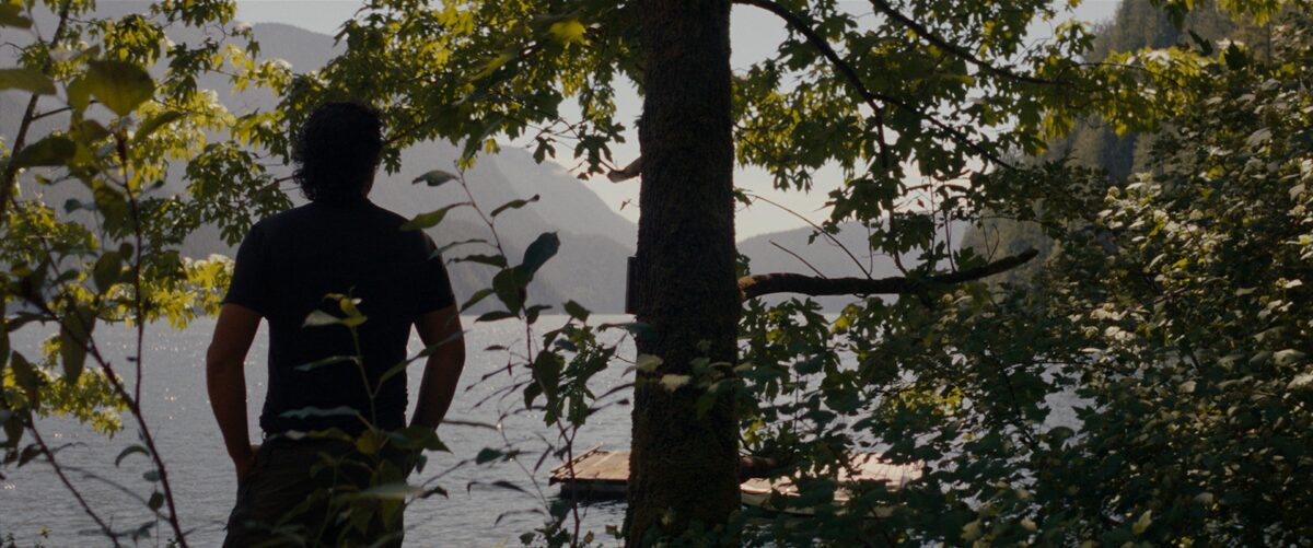 A person with dark hair wearing a black shirt standing on a lakeshore, surrounded by trees and foliage, with mountains and a dock in the background.