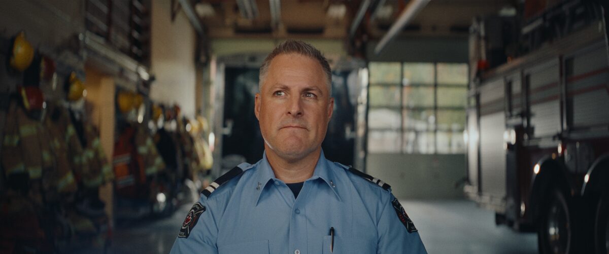 A male firefighter in uniform stands inside a fire station, looking to his left with a serious expression, surrounded by firefighting gear and equipment.
