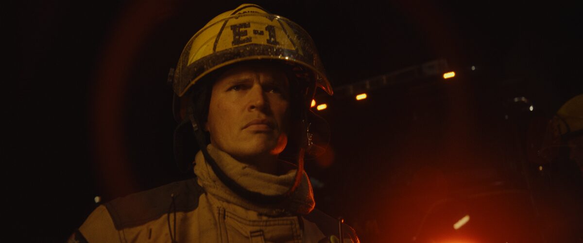 Close-up of a firefighter wearing helmet E-1, uniform, and protective gear, with a serious expression, illuminated by firelight in a dark environment.