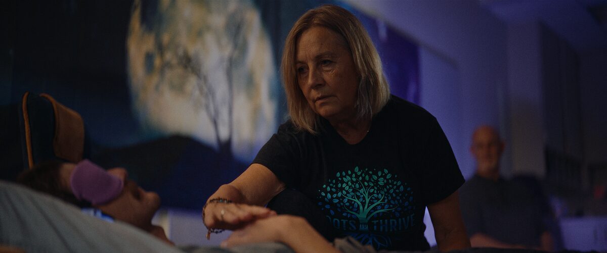An older woman with blonde hair, wearing a black T-shirt with a tree logo and the word 'Thrive,' is offering comfort to a person with a medical device on their forehead, lying in a care facility.